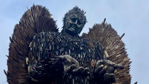 Looking up at the Knife Angel which is holding out its hands in front of its body. The structure is large with big wings. It has a sad facial expression. The sharp blades are poking out of the wings.