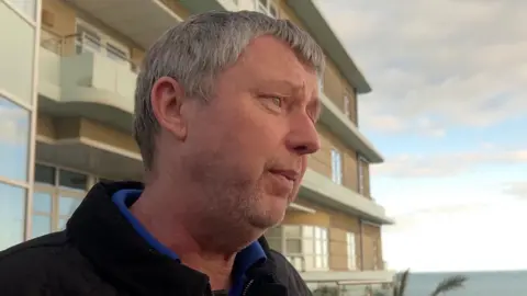 Andrew Wilkinson looking out of frame in a black jacket, with a blue shirt underneath, with the sea and a seaside hotel in the background.