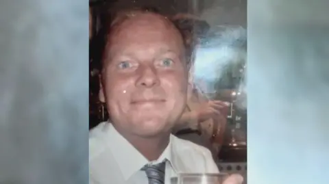Lee Hockey smiling at the camera. He is wearing a white shirt and a grey, black and silver tie. He is holding a drink in his right hand.