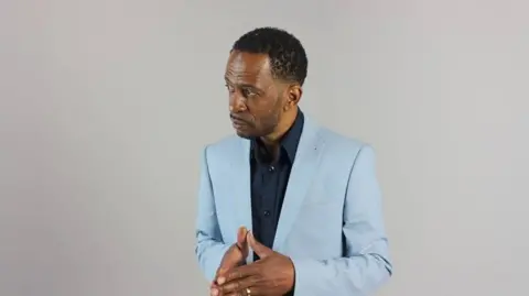 Granville Graham, wearing a light blue suit and navy shirt, in a studio head-and-shoulders photo in which he is looking slightly to the right. He has black hair and styled facial hair.