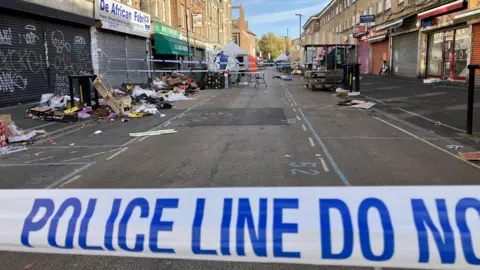 A close-up view of police tape across East Street, with buildings on either side of the road and market rubbish to both sides of the street