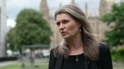 BBC The Labour MP for Truro and Falmouth, Jayne Kirkham pictured in front of the Houses of Parliament. She is wearing a black blazer and has light brown long hair. She is wearing an earring and a necklace.