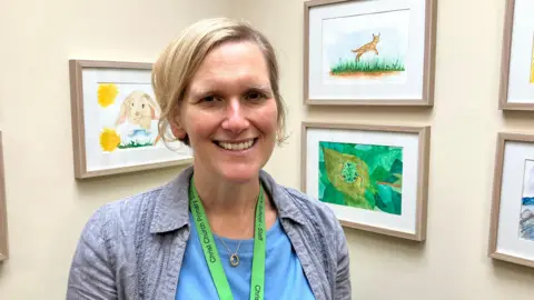 Headteacher Tracey Cansdale smiling in front of children's artwork inside her office. She is smiling at the camera and wearing a green lanyard round her neck.