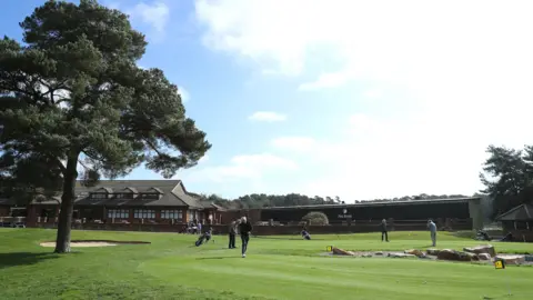 Getty Images Half a dozen people putting golf balls on a green in front of a large brown club house. There is a large tree on the left of the image