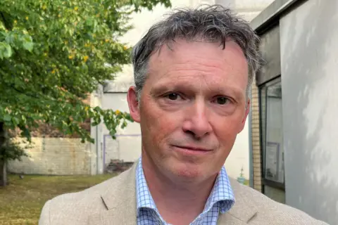 Close-up portrait of Richardson wearing a light-coloured blazer and blue checked shirt, standing outdoors beside a modern building. Trees and a courtyard area are visible in the background.