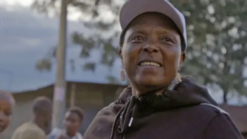 Menina, vestindo um tampo preto, boné branco e brincos de miçangas dourados, sorri enquanto entrega preservativos em uma rua em Naivasha.