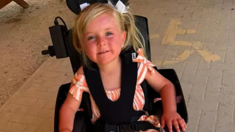 Warren Davies A young girl with blonde hair wearing a striped orange and white playsuit. She is sat in a black wheelchair with black straps holding her in. She is smiling and look at the camera.