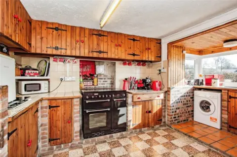 Zoopla/ John Francis A rustic kitchen with wooden cabinets, a black range cooker, tiled countertops, and a washing machine next to large corner windows.