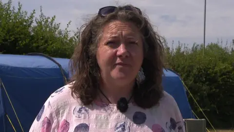 Nic Small/BBC A woman wearing a pink polka dotted shirt. She is stood in front of a blue tent, pitched in a tent.