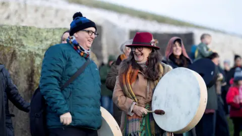 Reuters Two people holding drums. A crowd is gathered outside a stone structure with grass on its top.