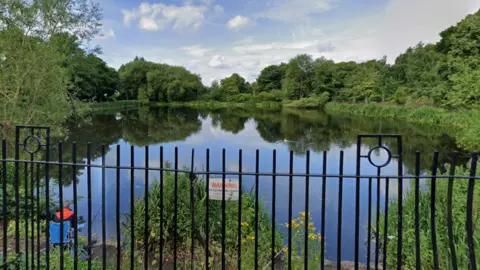 Google A reservoir with clear water. It is surrounded by numerous trees, and a fence with a warning sign on.