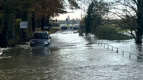 Harriet Heywood/BBC A road is submerged in water and a Range Rover is driving through, there is a sign pointing to the town centre and a church spire in the distance.