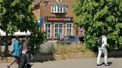 A shop with orange and brown Sainsbury's signage, with bushes in the foreground, and three people walking past.