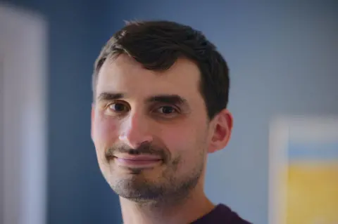 Cole's husband Ed is looking directly at the camera and smiling. The wall in the background is blue and there is a blue and yellow painting on the wall, blurred