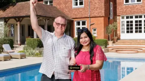 OMAZE David stands beside the pool of the new mansion he has won. He has his arm raised and is smiling. Behind him, the pool and part of the mansion.