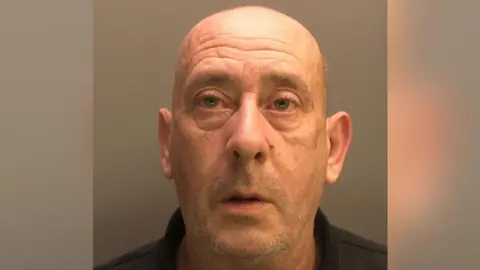 A police custody image of Mark Roberts, he has a bald head and stubble. He is wearing a black polo shirt and his mouth is slightly agape as he has his photo taken.