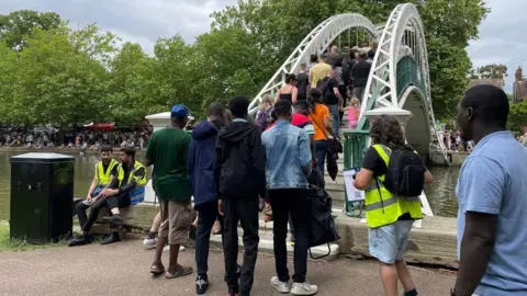 Bedford Borough Council A large group of people, walking over a bridge, forming a queue to get on it. Some people are sat down to the left. More people are seen in the distance on the other side of the river.