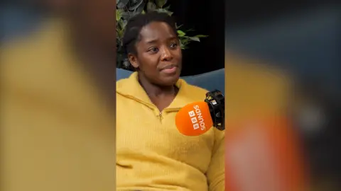 Kimberley Wilson, podcast host, is wearing a yellow T-shirt. She has black hair.