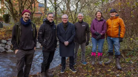 West Midlands Combined Authority Six people stand close to a small waterway and smiled at the camera. There are also trees behind the members of the group, who are all dressed in outdoor gear.