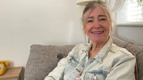 A woman smiling at the camera. She is wearing a denim jacket and is sat on a seat with part of a yellow telephone behind her. 
