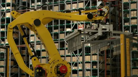 DJ McLaren/BBC A yellow robotic arm inside a warehouse. At the end of the arm is a grabber, which appears to be empty. Behind it are lots of shelves containing cardboard boxes. 
