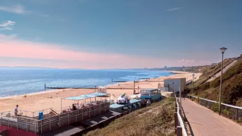 View from the zig-zag footpath path from the cliff top, looking down towards Sobo Beach bar and the long sandy beach of Southbourne and Bournemouth. The beach bar is made up of a converted double decker bus, a smaller building with air vents, and a large open decking area.