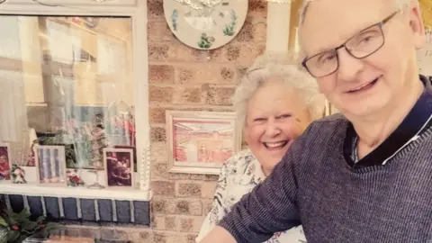 Hubert and Maureen Dodds smiling into the camera. Mr Dodds is standing slightly in front and to the right of his wife and looking slightly to the right. He is wearing a blue jumper and dark rectangular glasses. Mrs Dodds has short, curly white hair and is wearing a white patterned top. They are standing outside a brick house which has bits of artwork on the wall next to a window on the left.
