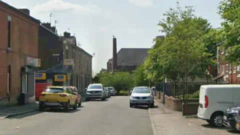 Google Google streetview of terraced houses and parked cars on Greenwood Street with trees to the right and a church in the backdrop.