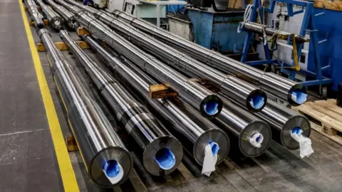 Eight long, shiny, metal tubes stacked on top of each other on a dark floor with blue machinery on the right hand side