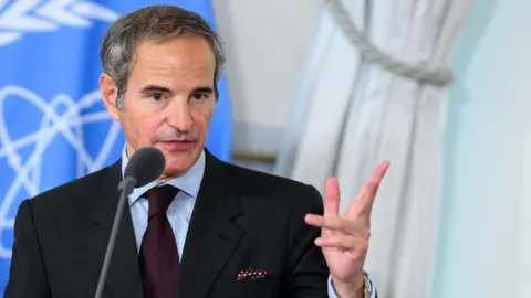 Rafael Grossi, gesturing with his hand and with a serious expression, speaking into a microphone. The blue IAEA flag hangs behind him. He has short grey hair and is wearing a black suit with blue striped shirt and maroon tie.