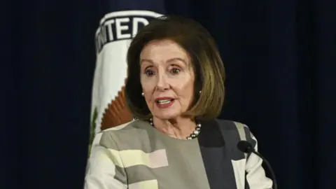 Nancy Pelosi delivers remarks at an award ceremony in Washington DC on 9 December. She has a brown bob hair-cut and wears a patterned blouse.