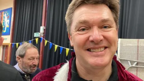 Jeremy Fish smiles at the camera inside a community hall. He has short dark hair and is wearing a red and white Santa Claus top. Behind him other volunteers at a wrapathon event can be seen talking.
