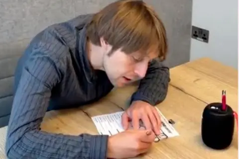 RNIB A man is filling in a ballot paper using a tactile voting aid, which is laid on top of the ballot paper, and an audio voting device which is placed next to him on a table. He has short, wavy brown hair and is wearing a blue striped shirt.