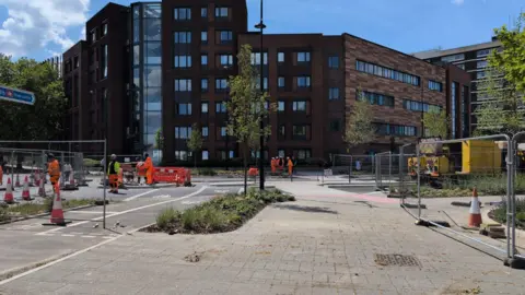 A foot way leads towards a multi-storey office building. The road next to the path is blocked off for road works, with people in orange high-vis working on it.