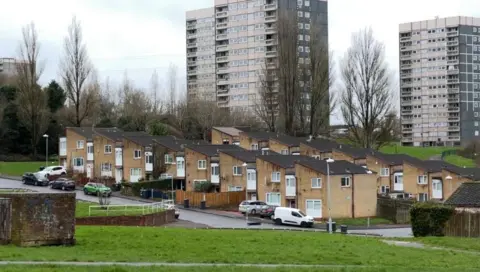 BBC Tower blocks are seen rising up behind a row of houses at Druids heath. There are open spaces with grass and trees dotted around the landscape. Cars are parked in the road outside the houses.