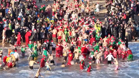 A crowd of people, many wearing festive, red hats, wade in the sea.