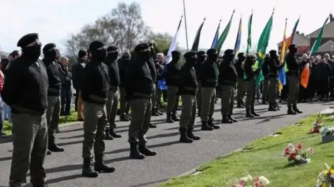 PA Media A number of masked men wearing berets , combat trousers and black jumpers stand at a commemoration holding Irish nationalist flags