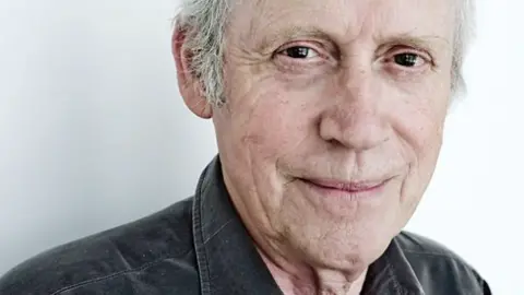 Archie Fisher is looking into the camera and half smiling. He has grey hair but the photo has been cropped at his forehead. He is wearing a grey shirt.