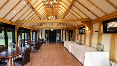 Matt Wing/Google The inside of one of the buildings at The Old Essex Barn. There is an open beamed roof with yellow wooden beams and white ceiling panels. To the right is a long table with a white tablecloth on top, and to the left are several round tables and chairs, and there is a tiled floor.