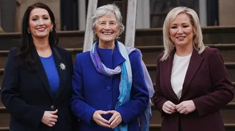 PA Media Catherine Connolly, centre, with short grey hair, wearing a blue coat. Deputy First Minister Emma Little-Pengelly is standing to her left, wearing a blue top and a navy jacket. First Minister Michelle O'Neill is standing to Connolly's right, wearing a white top and a burgundy jacket. 