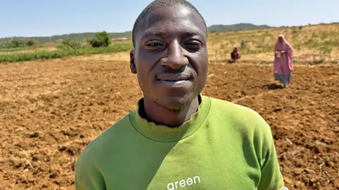 BBC/Alex Last Amos, a Berom farmer, in a light green sweatshirt with the word 'green' printed in white looks at the camera. Behind him is a ploughed field and two women out of focus.
