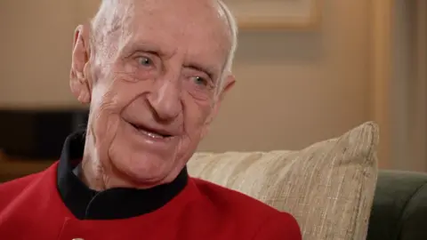 An elderly gentleman wearing a scarlet jacket smiles