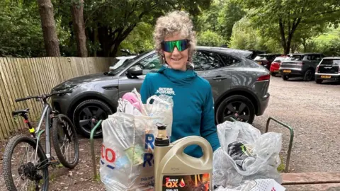 Lindsay Buck Lindsay Buck standing behind a wooden bench with a collection of rubbish. It includes a full Tesco carrier bag, a large plastic tub and a plastic bag with empty tin cans. She has grey hair and is wearing reflective sunglasses and a blue top. A bike is positioned next to her and a number of parked cars can be seen behind her.
