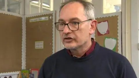 A mean wearing a blue round-neck jumper over the top of a red shirt is talking into a camera in a school classroom setting. He has receding grey hair and black-rimmed glasses.