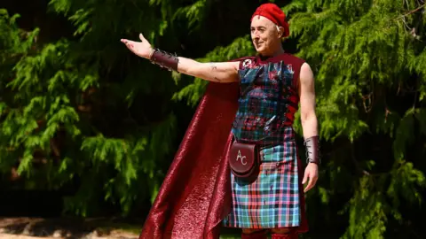 Alan Cumming, a man, is stood holding his arm out with trees behind him. He has a red beret on his head, a leather wrist band, a red cape, a tartan vest, tartan kilt, red knee high socks and a sporran which have the initials AC embroidered onto them. He is smirking slightly and looks as if he is welcoming people to something.