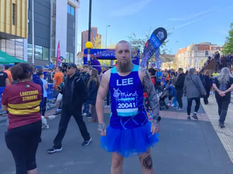 Lee McGhie is wearing a light and dark blue top with Lee and the logo of mental health charity Mind on. He has a beard and is also wearing a blue tutu