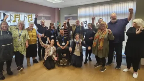 Zeb Soanes In a room with a wooden floor, fifteen people, including a man in a wheelchair, raise their arms in celebration
