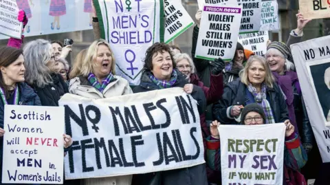 PA Media An image of a protest by the group Women Won't Wheesht, featuring a group of women holding signs with mottos like "no males in female jails", "violent males still in women's jails", "Scottish women will never accept men in women's jails", and "not men's shield".