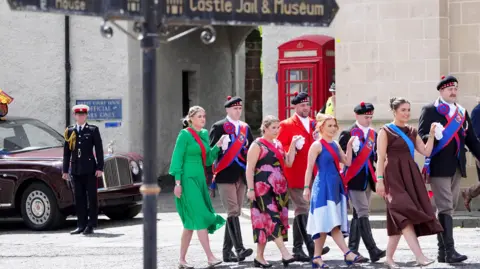 PA Media A group of men and women in colourful dress walk hand in hand through Jedburgh