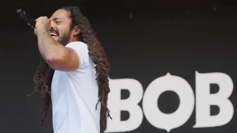 Bob Vylan frontman Bobby Vylan performing at Glastonbury Festival. He is wearing white polo shirt and has several tattoos on his arms. The word 'Bob' is behind him on the stage.
